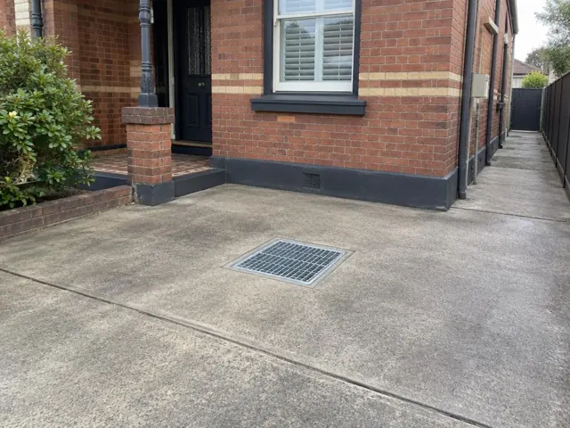 Clean outdoor gully grate set in dry concrete paving beside a brick house in The Hill